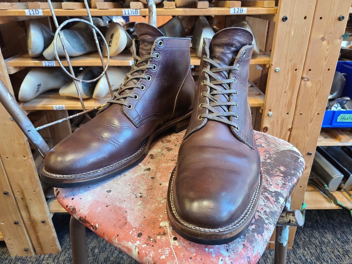 Pair of brown leather work boots on a pink, paint-splattered workbench, with shelves of shoe lasts and a metal shoehorn in the background.