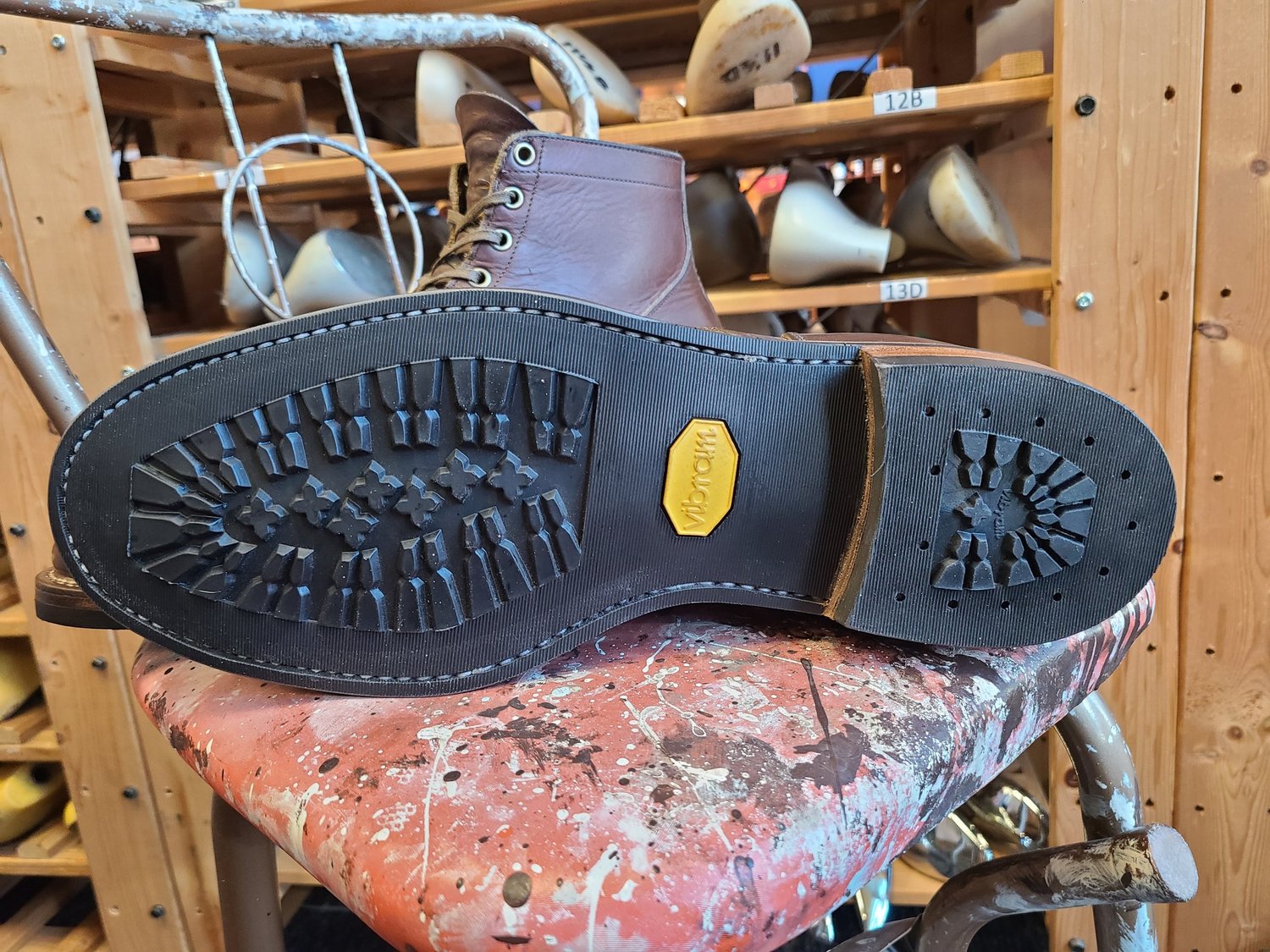 Close-up of a brown leather boot displaying the sole with a yellow Vibram logo on a painted, splattered stool. Shelves in the background hold ceramic mugs.