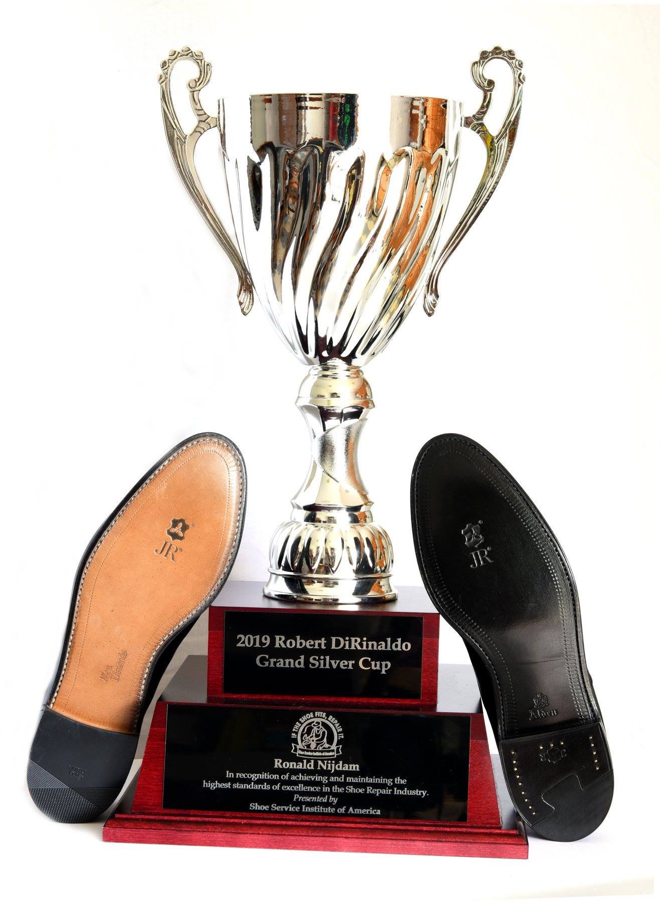 A large silver trophy cup with ornate handles, placed on a red base with two plaques. The plaque reads '2019 Robert DiRinaldo Grand Silver Cup'. In front of the trophy are two shoe soles, one tan and one black, with inscribed logos.